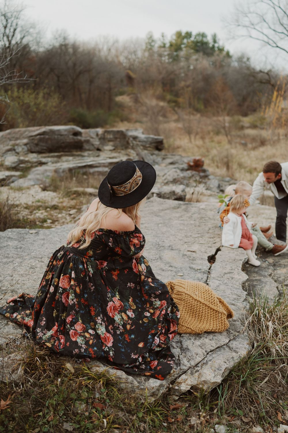 Woman in floral dress and black hat sits on rocks watching two young children play in an autumn outdoor setting.