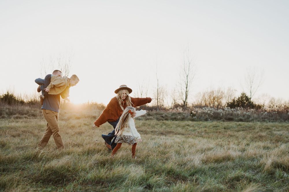 Family playing in a sunlit field at golden hour, parents twirling children in a rustic outdoor setting.