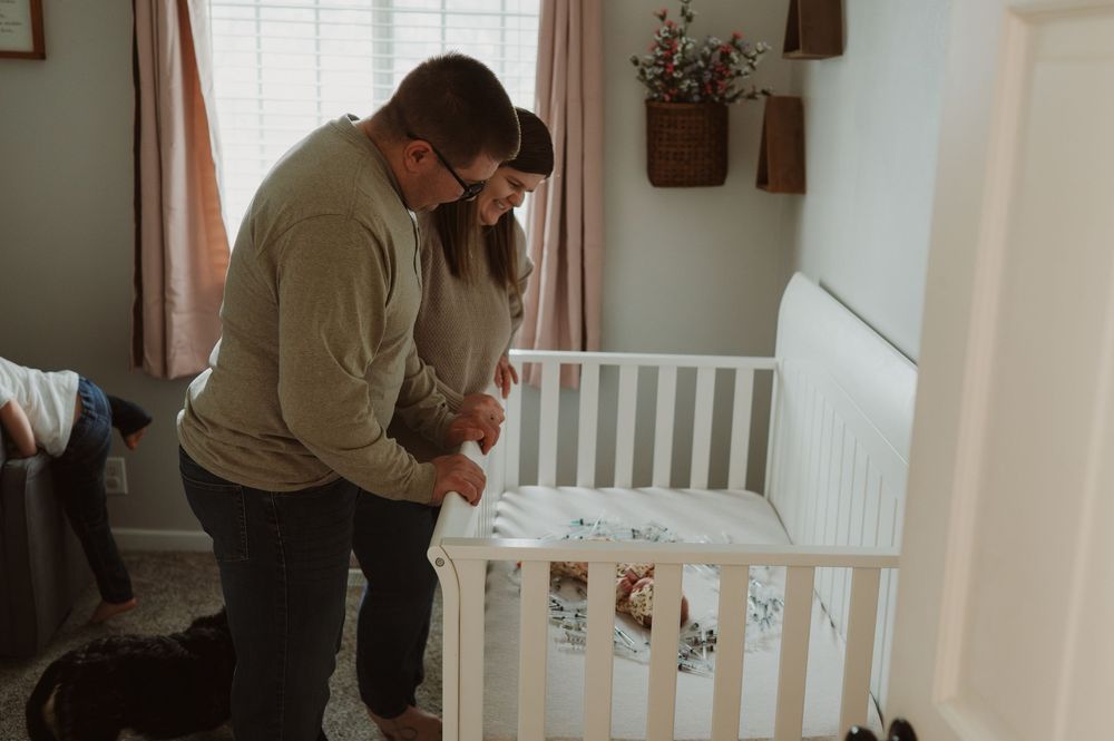 A couple lovingly gazes at their newborn baby lying in a white crib in a softly lit nursery.