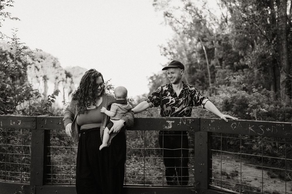 Black and white photo of a couple with their baby on a wooden bridge surrounded by lush trees.
