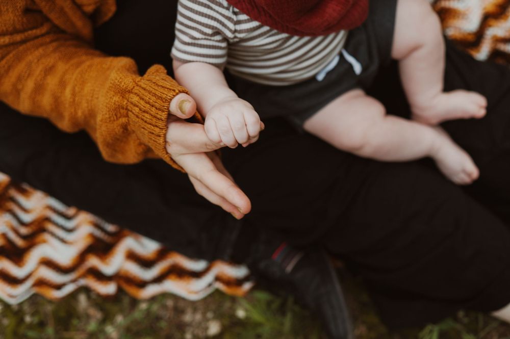 A baby grasping an adult's finger while sitting on a lap, wearing a striped outfit with a colorful blanket nearby.