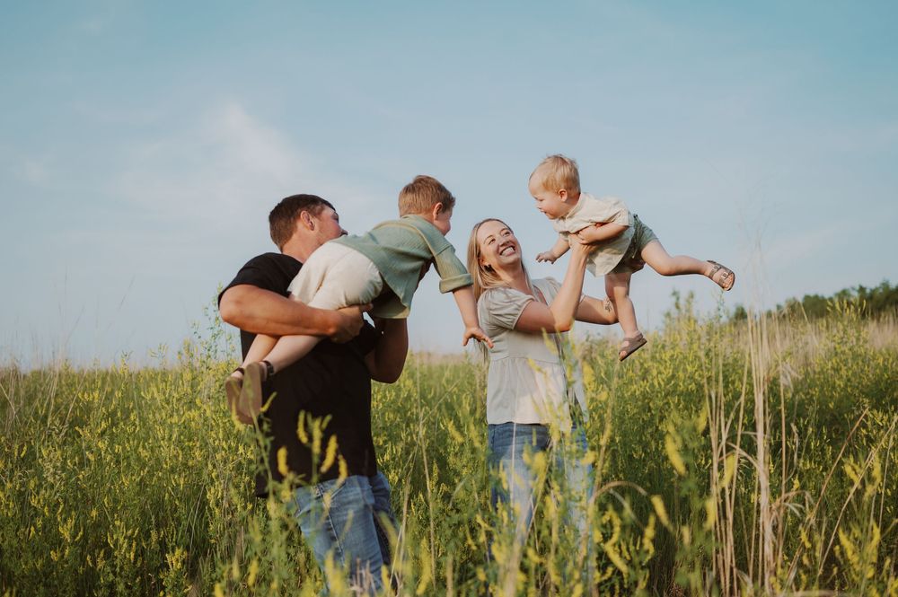 Family of four playing in green field, parents lifting two young boys in the air on sunny day.