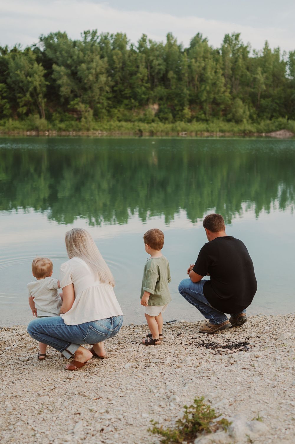 Family of four crouching by a serene turquoise lake surrounded by green trees.