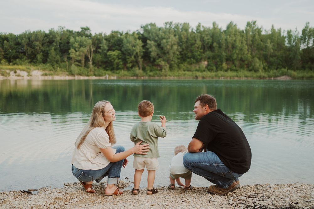 Family of three crouching by a serene blue lake shore, sharing a peaceful moment together outdoors.