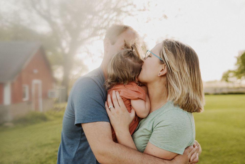 A couple shares a tender kiss while embracing a child outdoors near a red brick house on a sunny day.