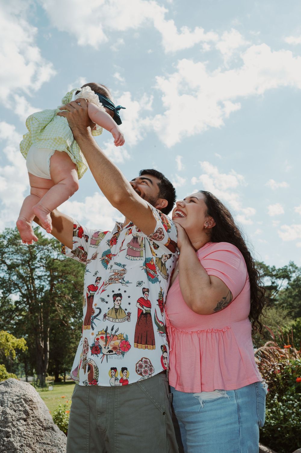Parents in casual attire lift and play with a baby outdoors against a blue sky and trees on a sunny day.