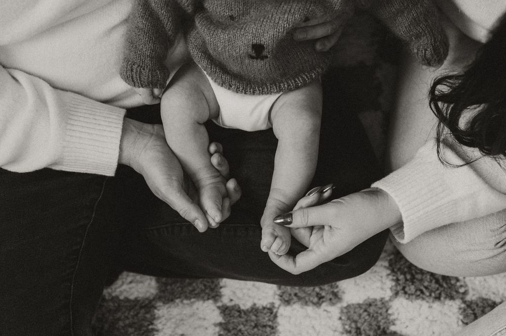 Black and white photo of parents holding their newborn baby's tiny feet and hands on a checkered blanket.