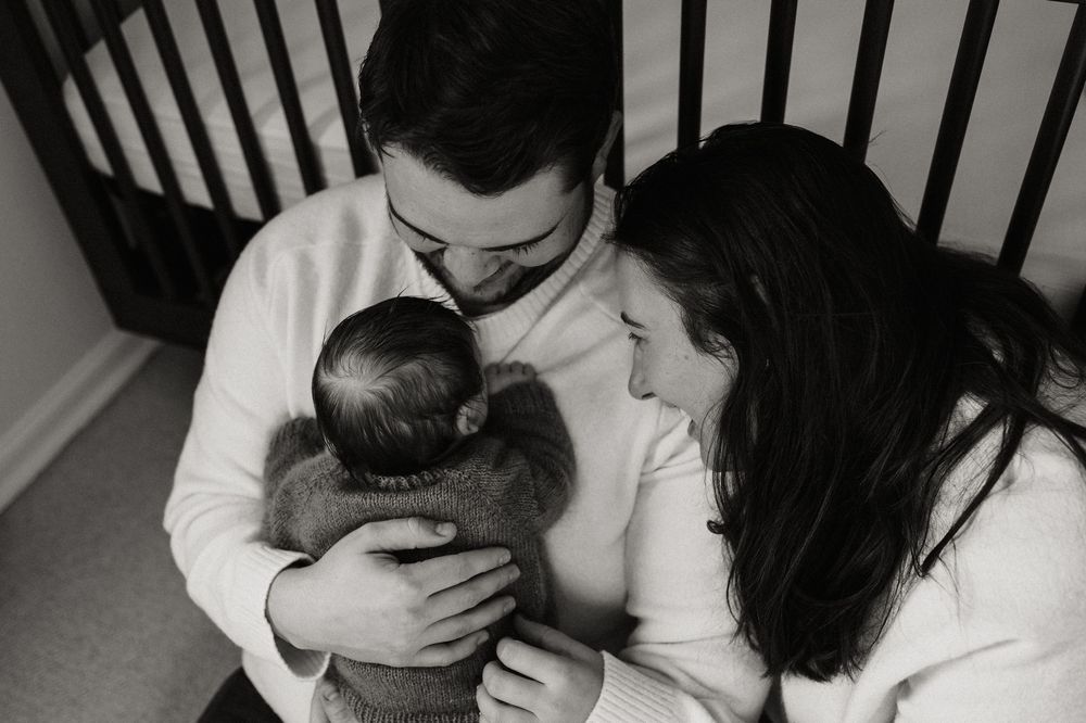 Black and white photo of parents lovingly cradling their newborn baby together indoors.