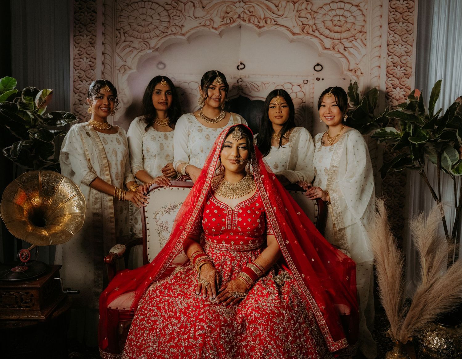 Bridal party in white outfits surrounding bride in red lehenga against decorative backdrop.