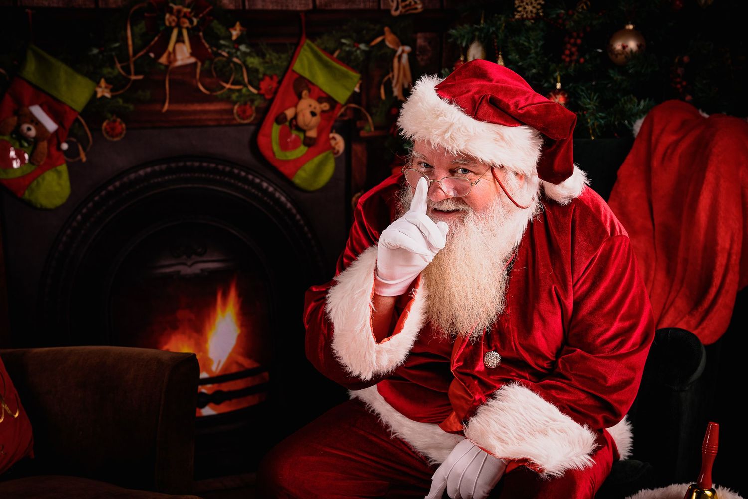 Santa Claus sitting by fireplace with finger to lips in Christmas setting.