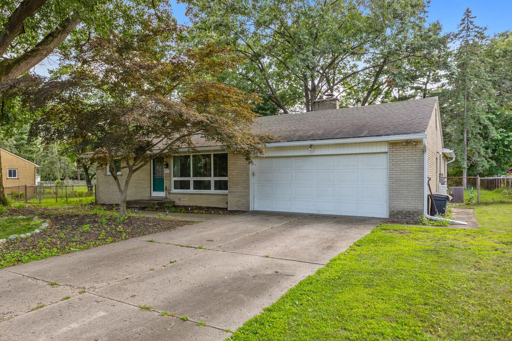 Single-story ranch home featuring a white garage door, mature trees, and well-maintained green lawn.