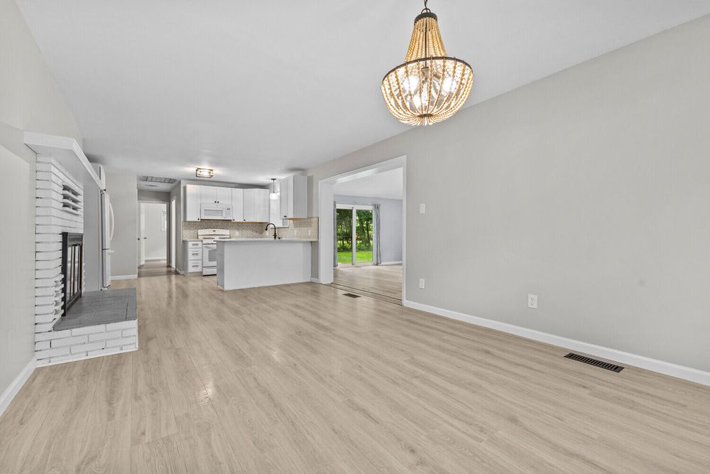 Open-concept living room with light hardwood floors, brick fireplace, chandelier, and view of kitchen.