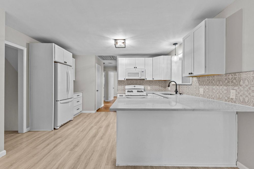 Modern white kitchen with peninsula, white appliances, decorative backsplash, and light wood floors.