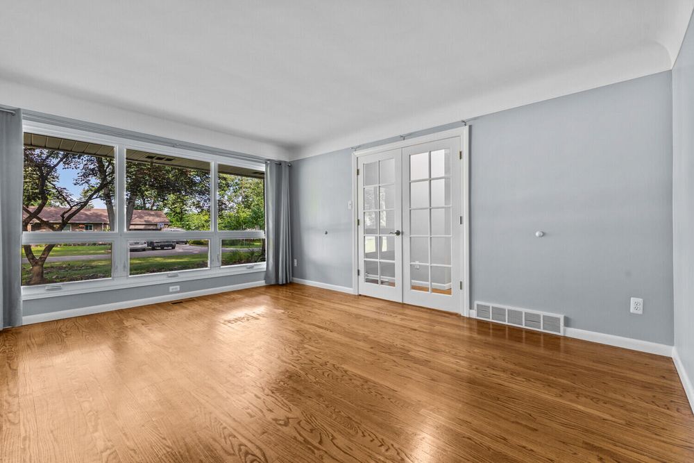 Sunlit empty room with hardwood floors, French doors, crown molding, and large windows with garden view.