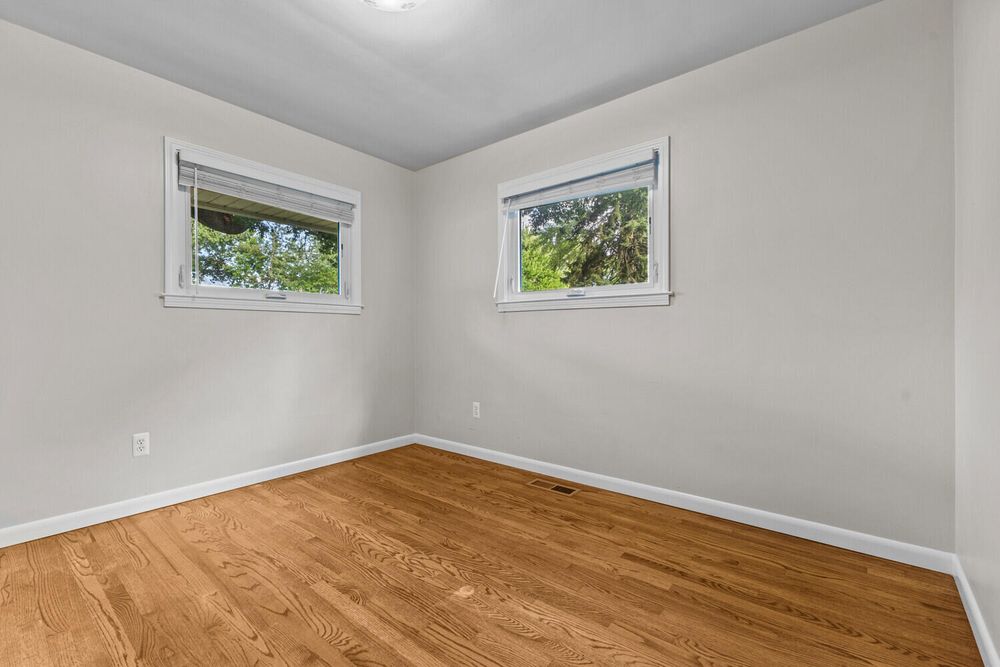 Empty bedroom with two windows, light hardwood floors, and neutral light gray walls.