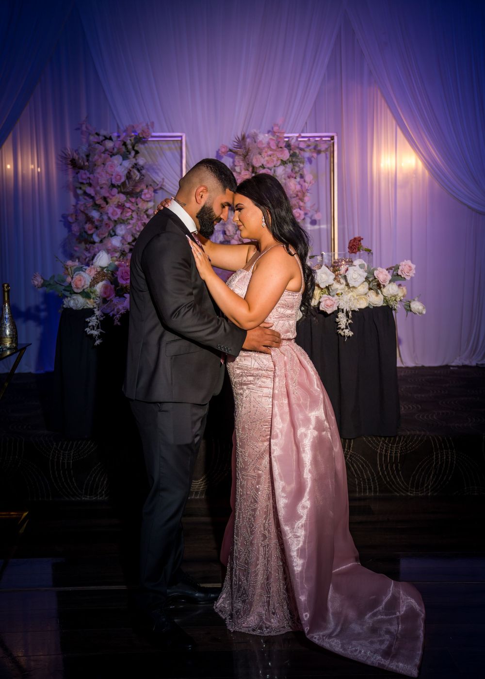 A romantic moment captured under purple lighting with floral decorations and draped fabric.
