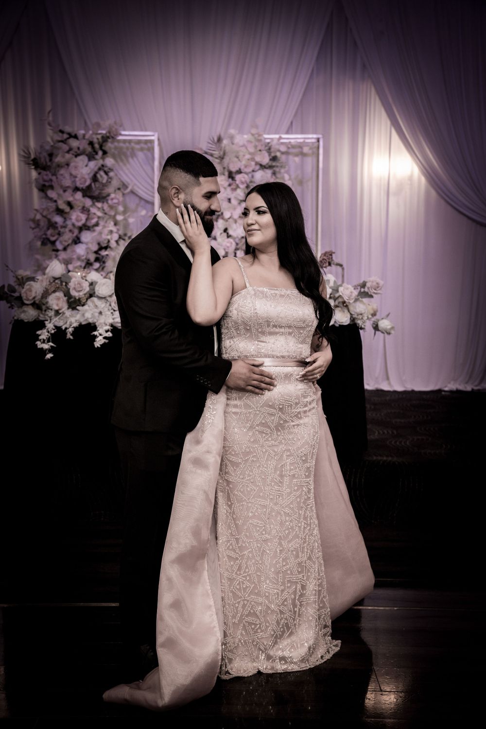 Romantic moment between newlyweds against purple-lit floral backdrop at reception venue.