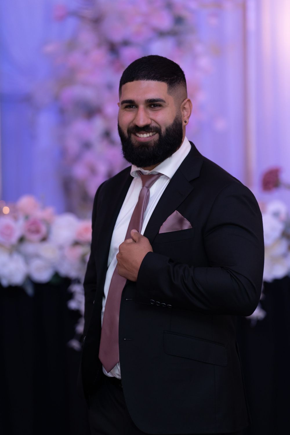 A groom in classic black suit and pink tie smiles against a purple-lit floral backdrop.