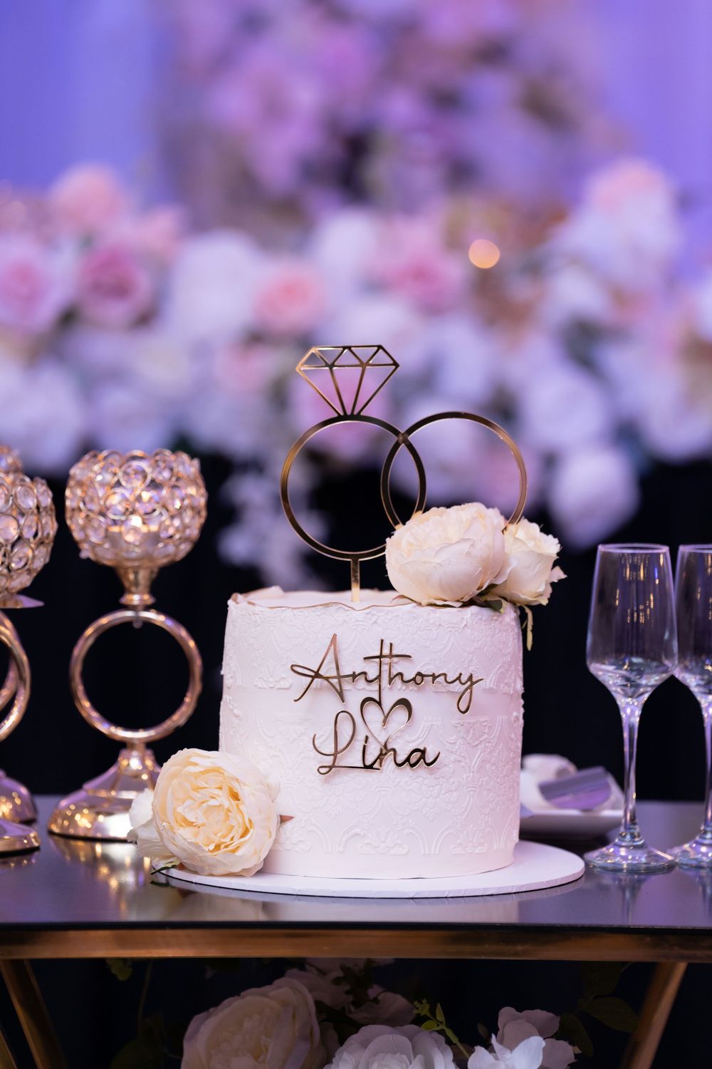 A white wedding cake with ring topper and floral decorations displayed against a purple backdrop.