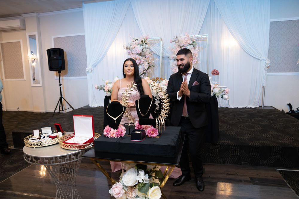 Couple celebrates at wedding reception opening gifts and making toasts with ornate floral backdrop.