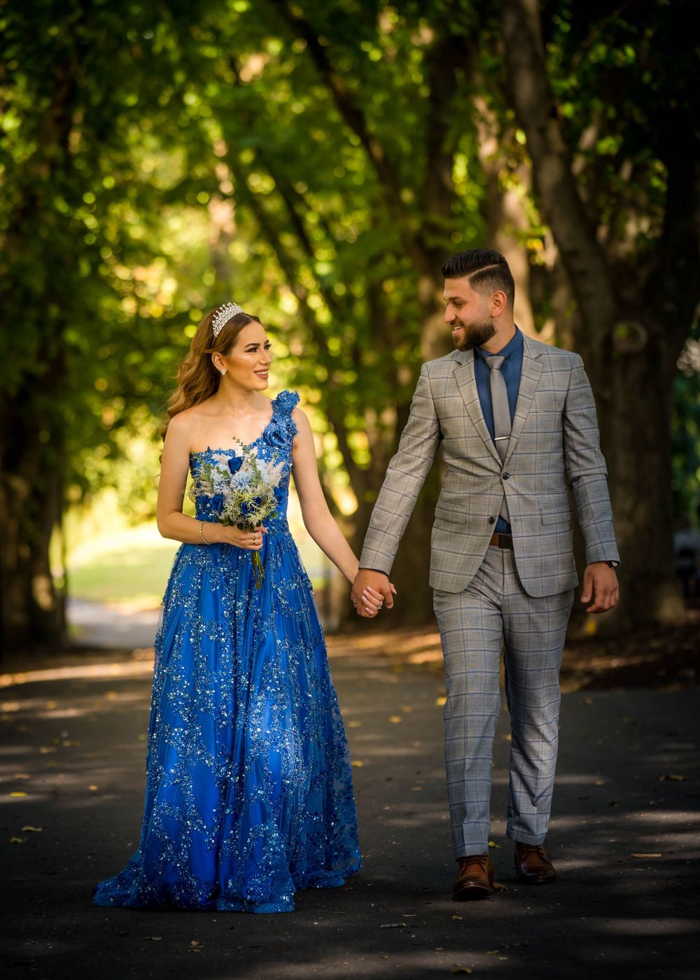 Wedding couple strolling down tree-lined path in blue gown and grey suit.