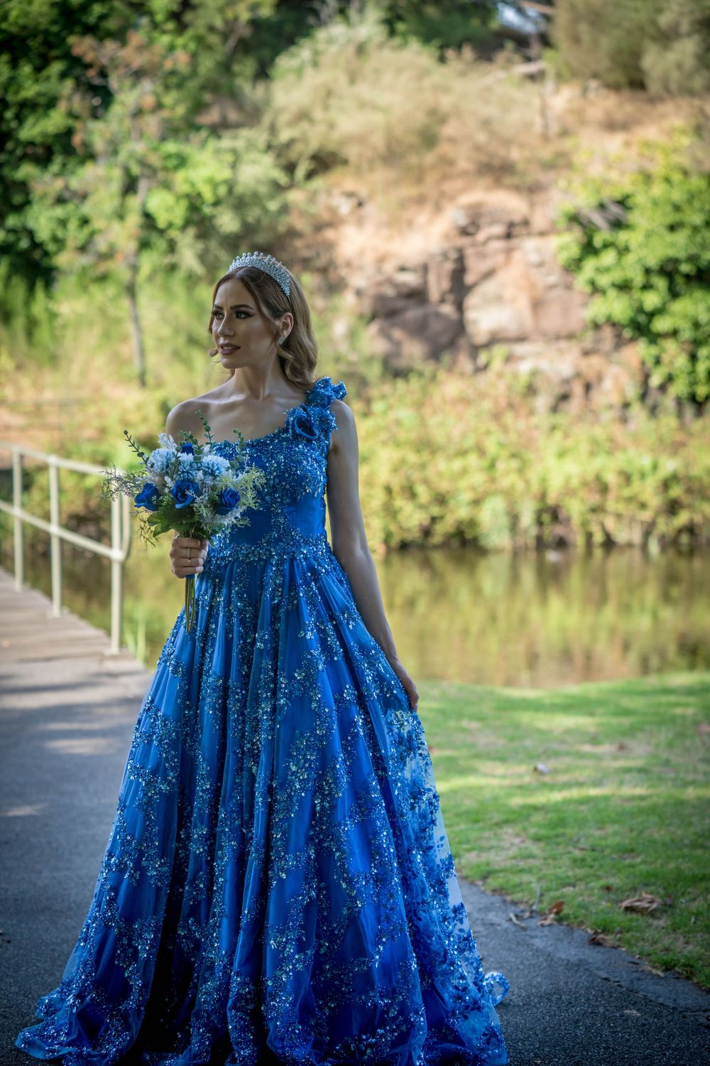 Woman in a sparkling blue ball gown holding a bouquet on a lakeside path.