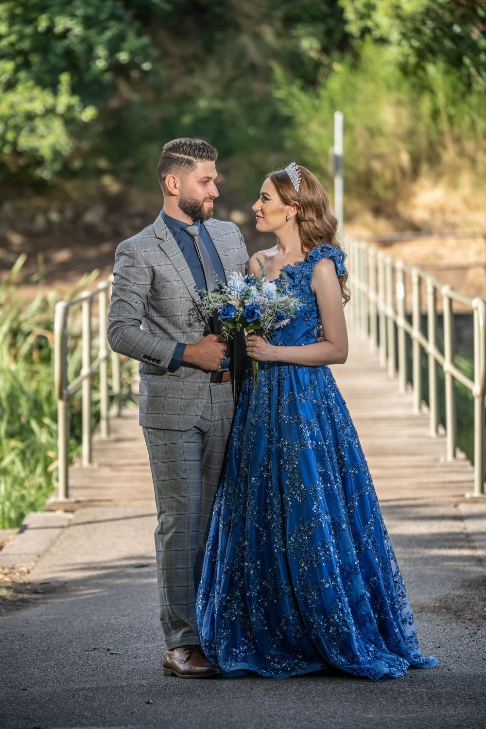 Couple sharing a moment on a bridge pathway, she in blue gown and he in gray suit holding flowers.