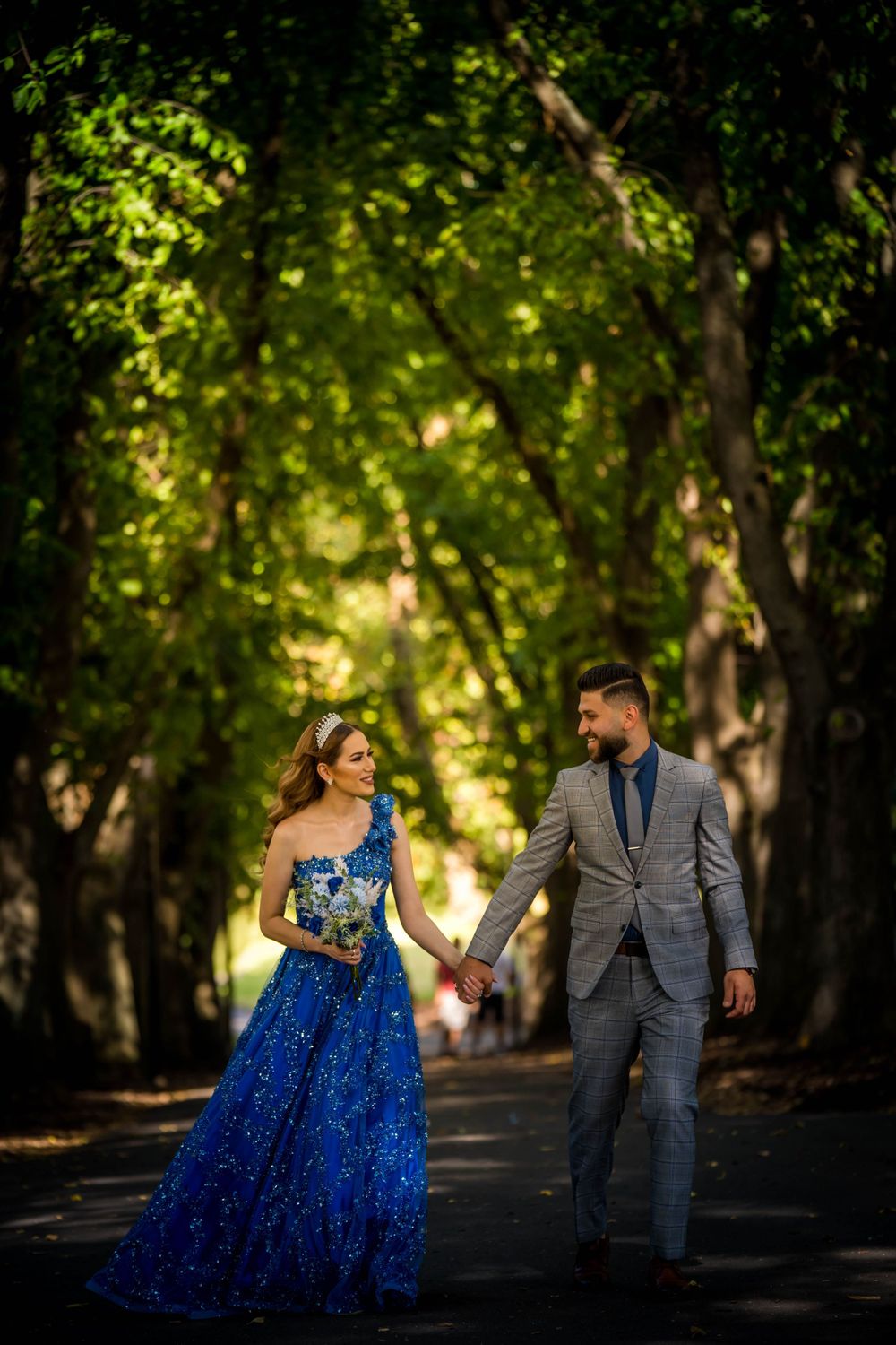 Couple in formal attire holding hands while walking through sunlit forest path.