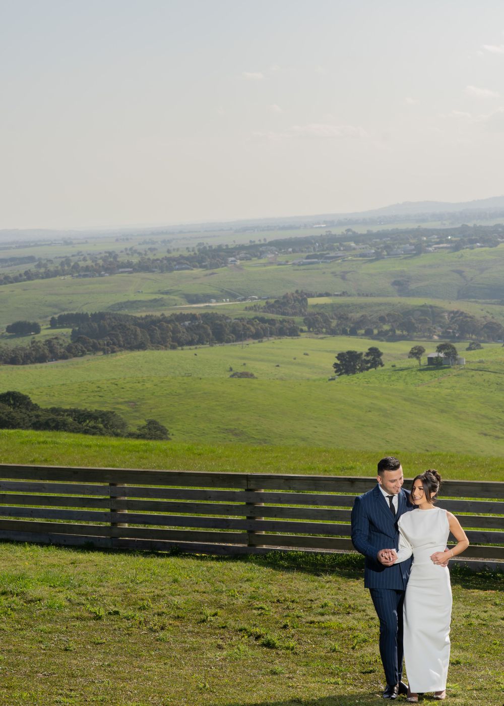 Newlyweds stand together by rustic fence with panoramic views of green pastoral landscape.
