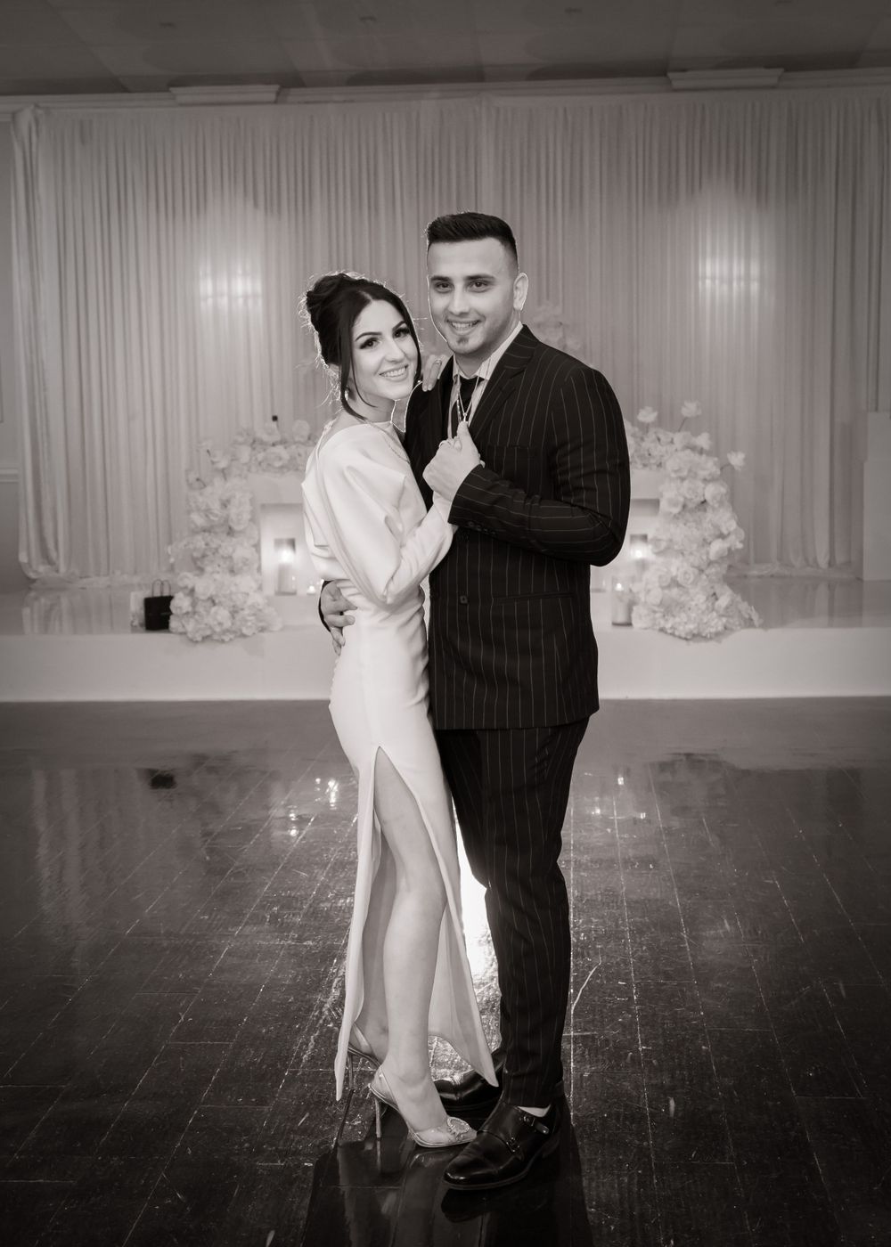 Elegant couple poses in formal wear against white floral backdrop at a glamorous event.