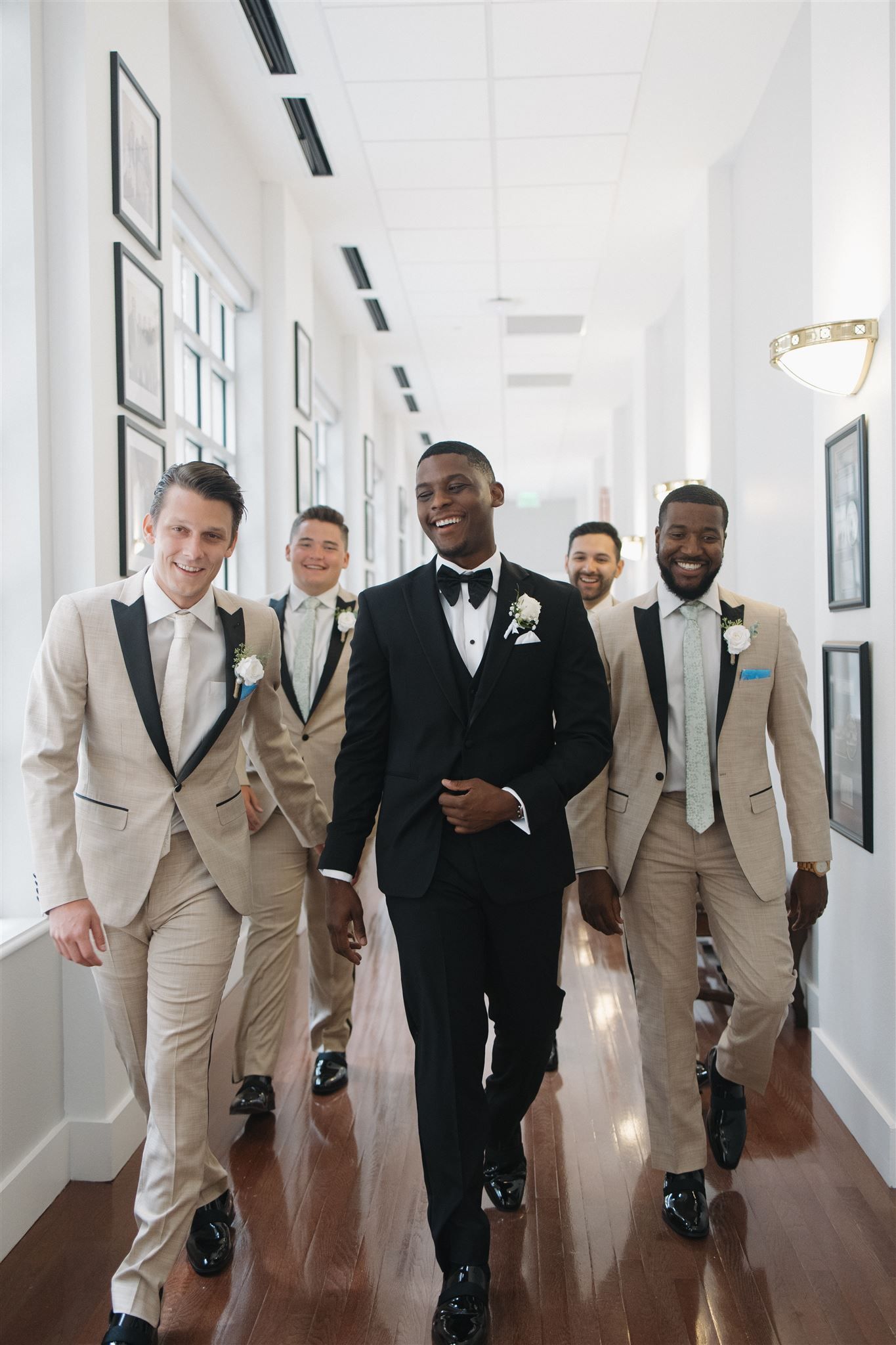 Wedding party wearing beige suits walking down a bright hallway with black and white framed photos on the walls.