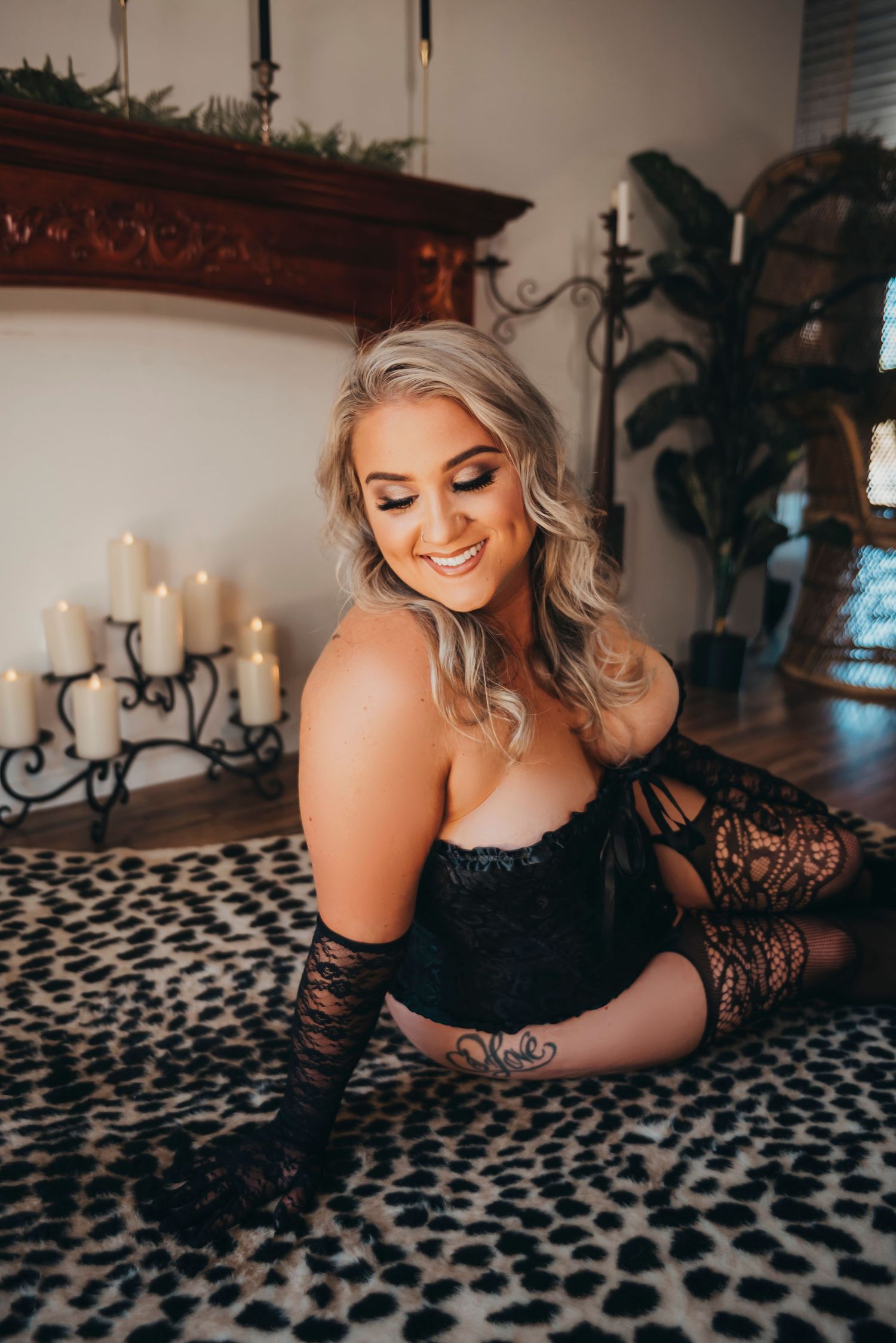 Woman in black lingerie, smiling playfully while kneeling on a leopard print rug, surrounded by candles, showcasing confidence and beauty.
