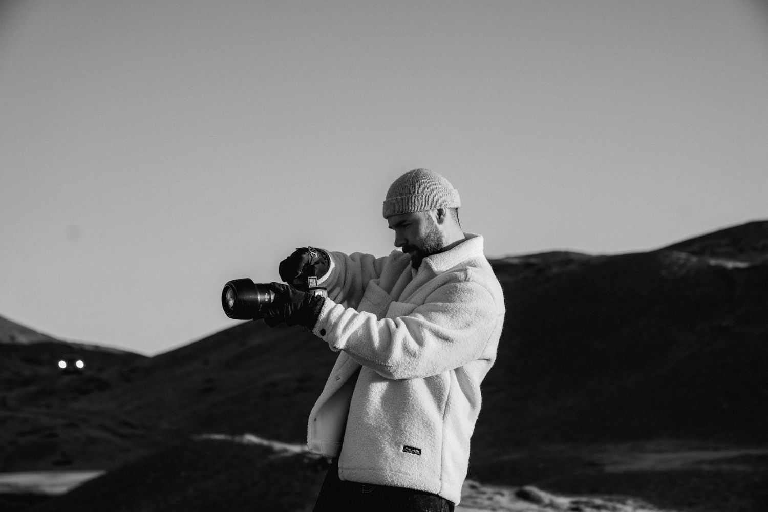 A photographer in a white winter coat takes aim with a camera against a mountainous landscape in black and white.