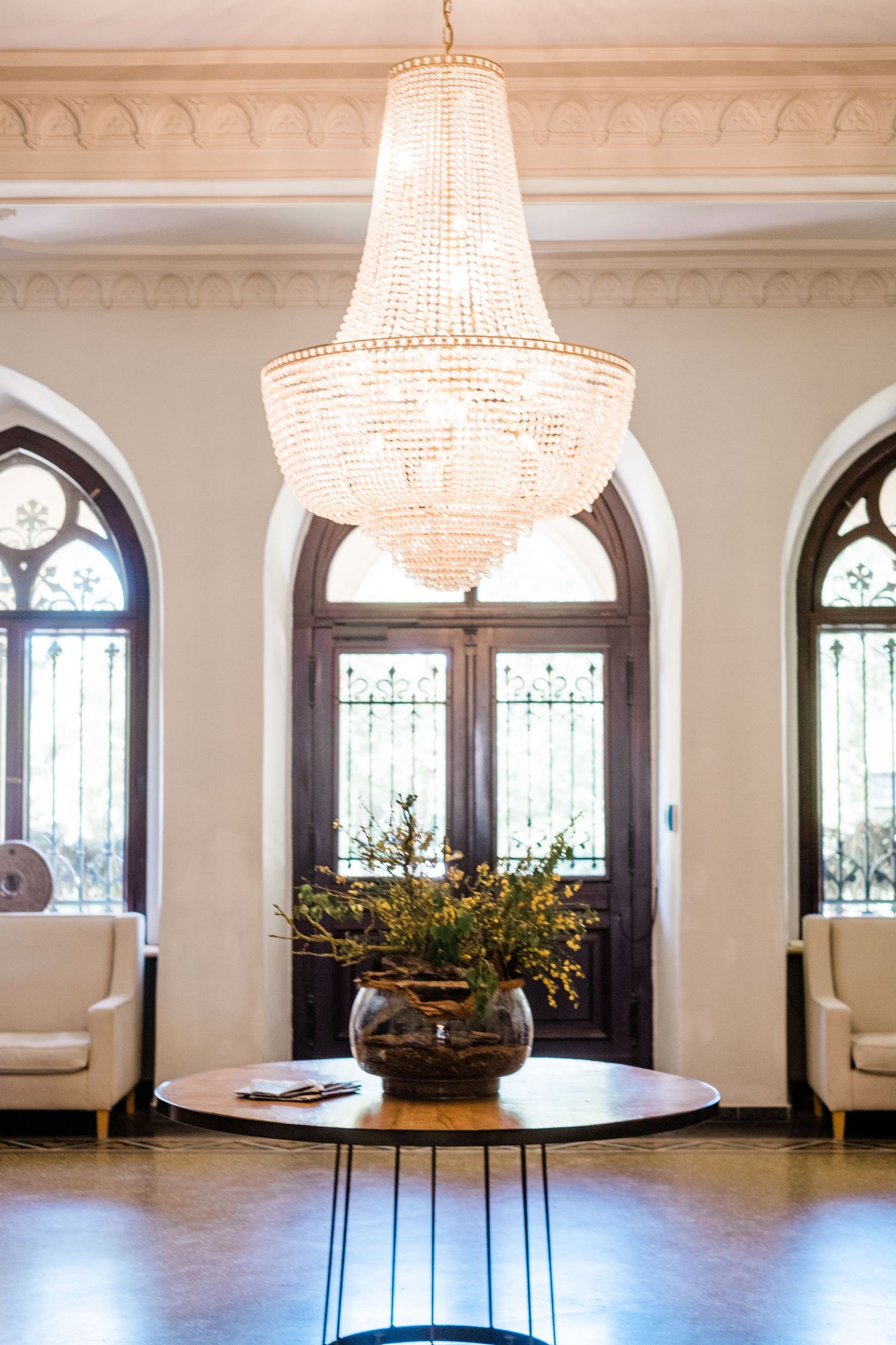 Grand crystal chandelier hangs above round table in elegant room with arched windows and cream-colored walls.
