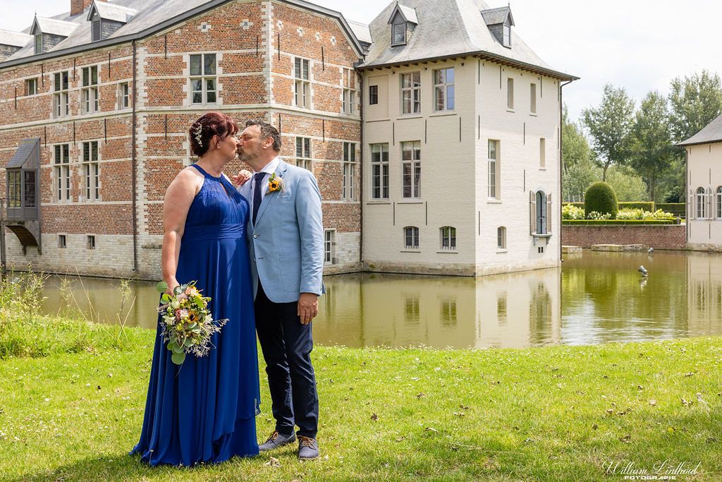 A couple in formal attire poses by a historic castle with reflective pond in background.