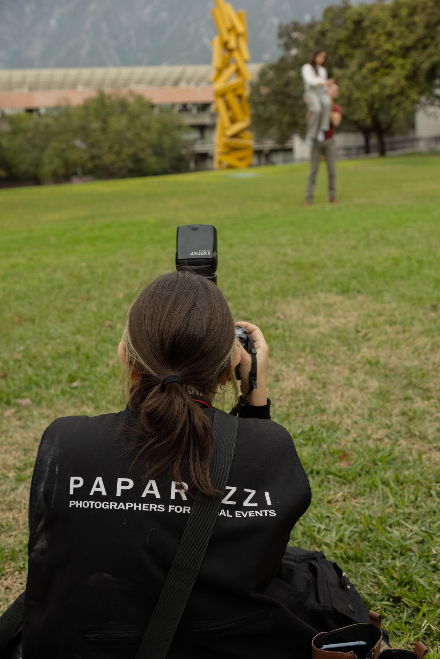 Paparazzi photographer sitting on grass, photographing a subject near a yellow sculpture outdoors.