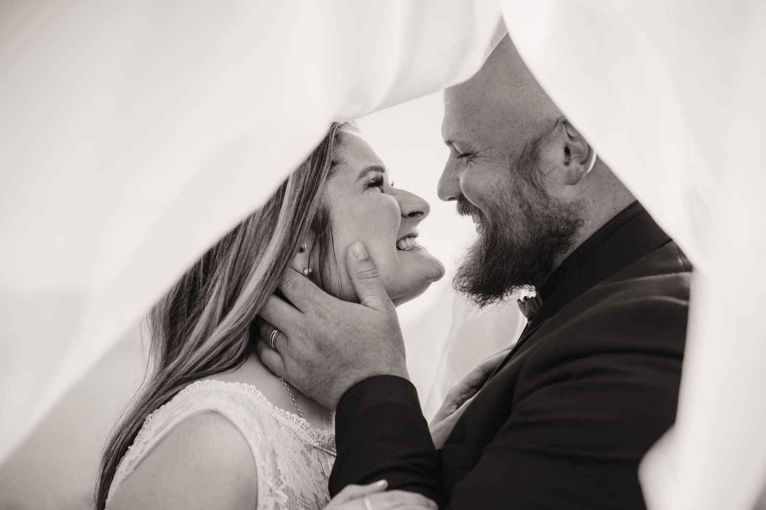 Black and white romantic wedding photo of couple laughing together while holding each other under a veil.
