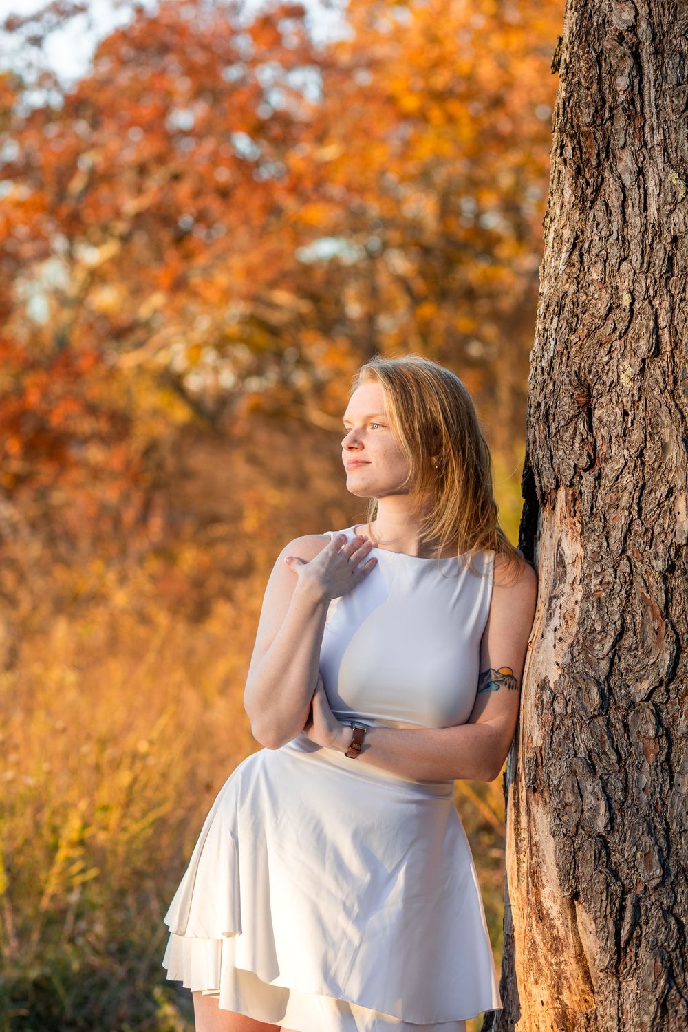Young woman in white dress leaning against tree trunk in autumn forest with golden orange foliage background.