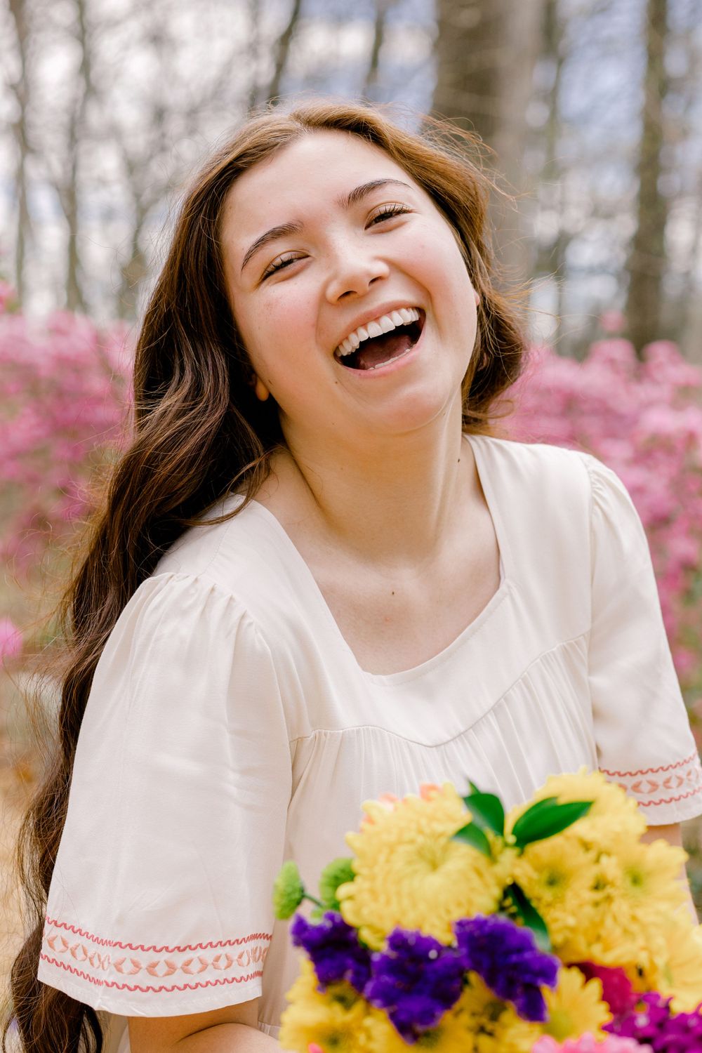 Person laughing joyfully while holding colorful flowers in a spring garden setting with pink blossoms in background.