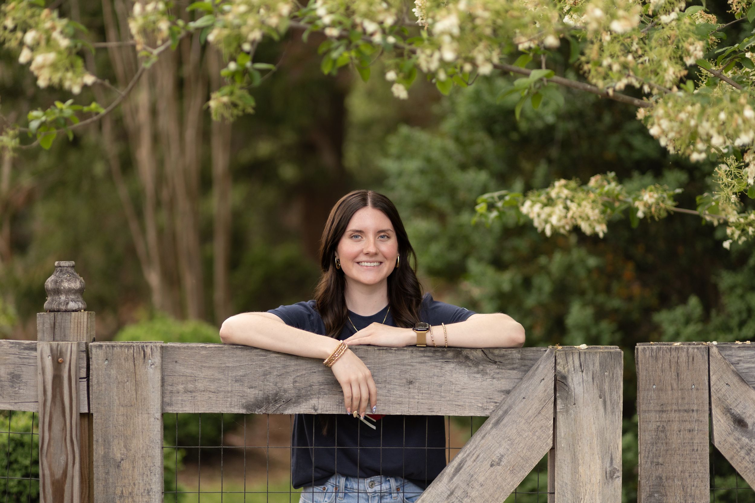 Young woman smiling and leaning on a rustic wooden fence surrounded by lush green trees outdoors.