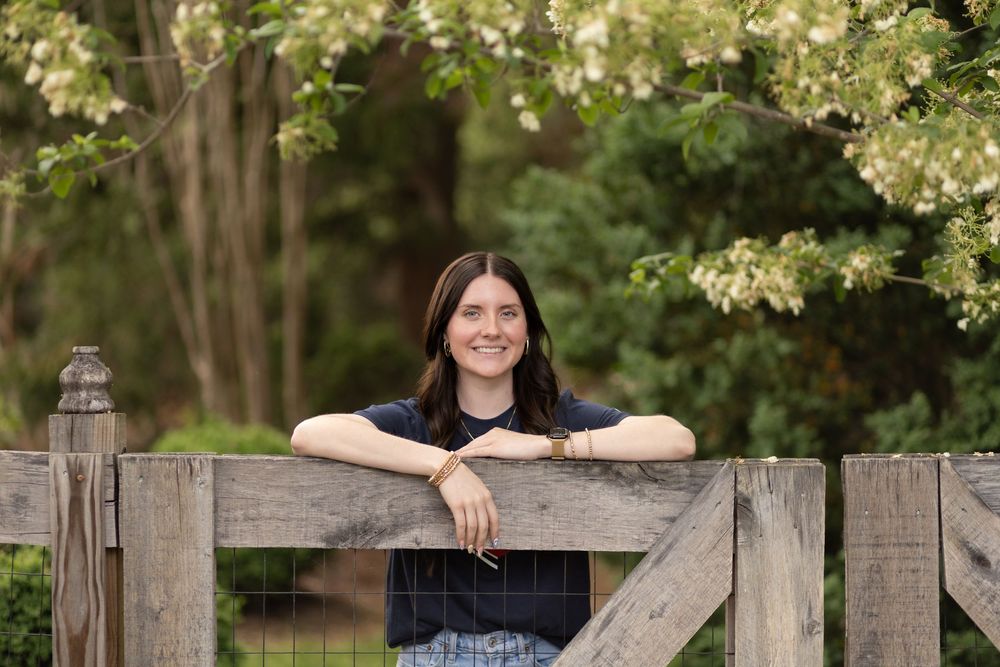 Young woman smiling and leaning on a rustic wooden fence surrounded by lush green trees outdoors.
