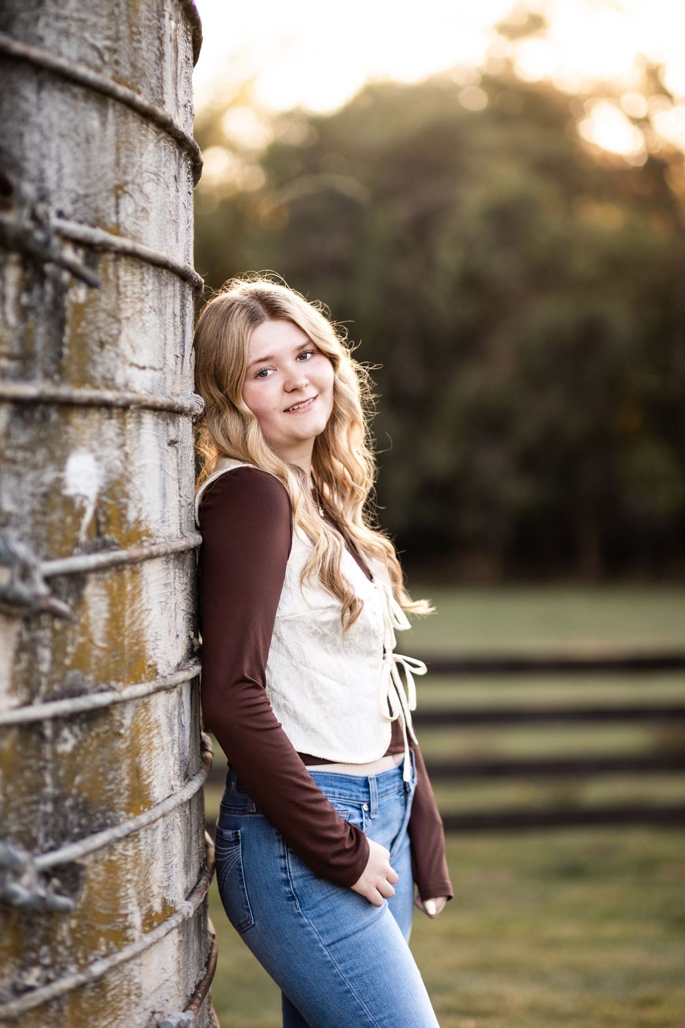 Young blonde woman in jeans and maroon top leaning against rustic silo at golden hour on a farm.