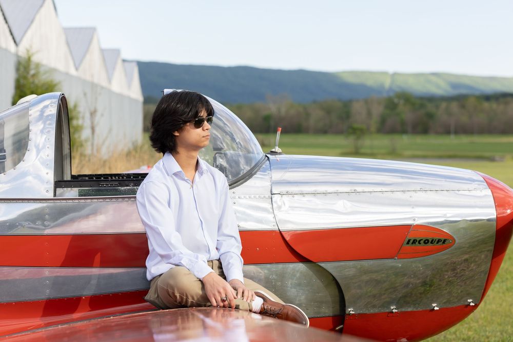 Young man in sunglasses leaning on a red and silver small aircraft on a green airfield with hills in background.