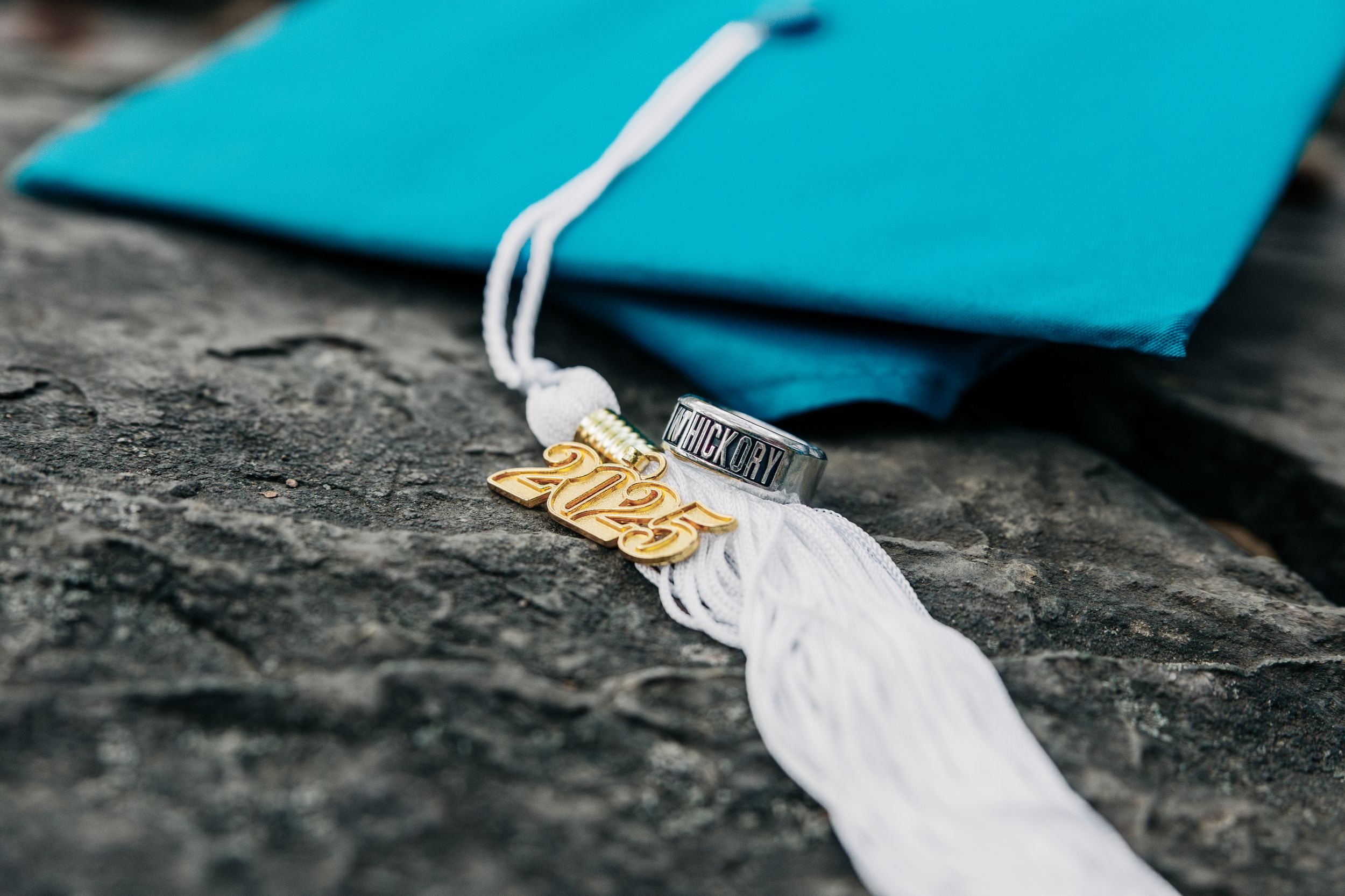 Graduation tassel with white and gold colors resting on a dark stone surface next to a turquoise colored item.