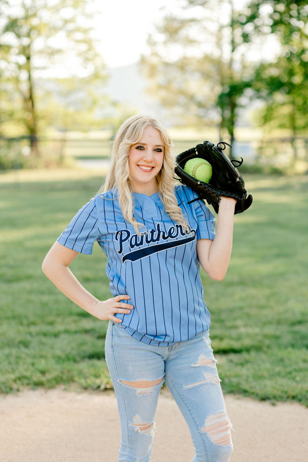Person in blue Panthers baseball jersey and ripped jeans poses with softball glove in sunny outdoor setting.