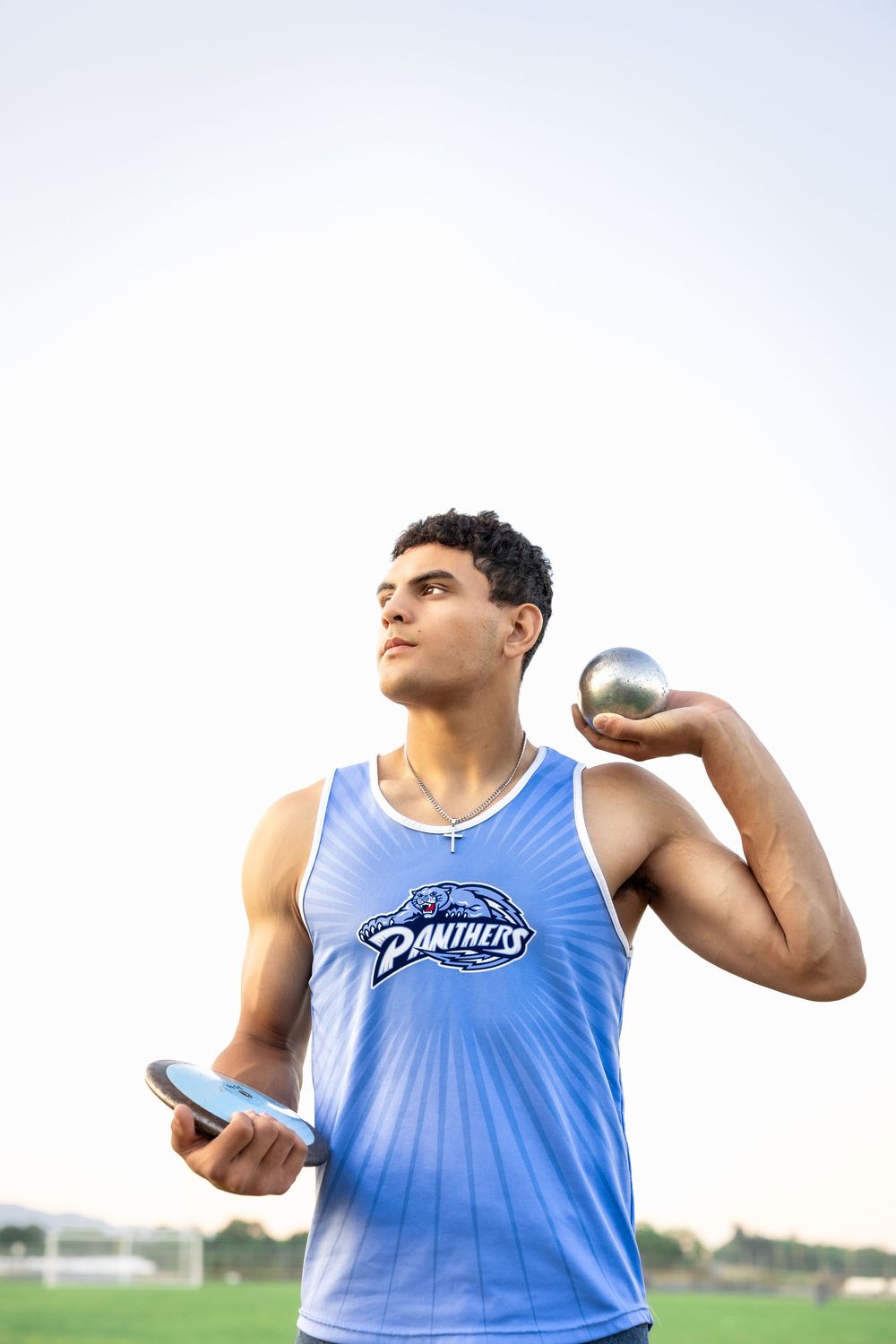 Athlete in blue Panthers tank top holds shot put ball and phone while practicing at sports field during sunset.