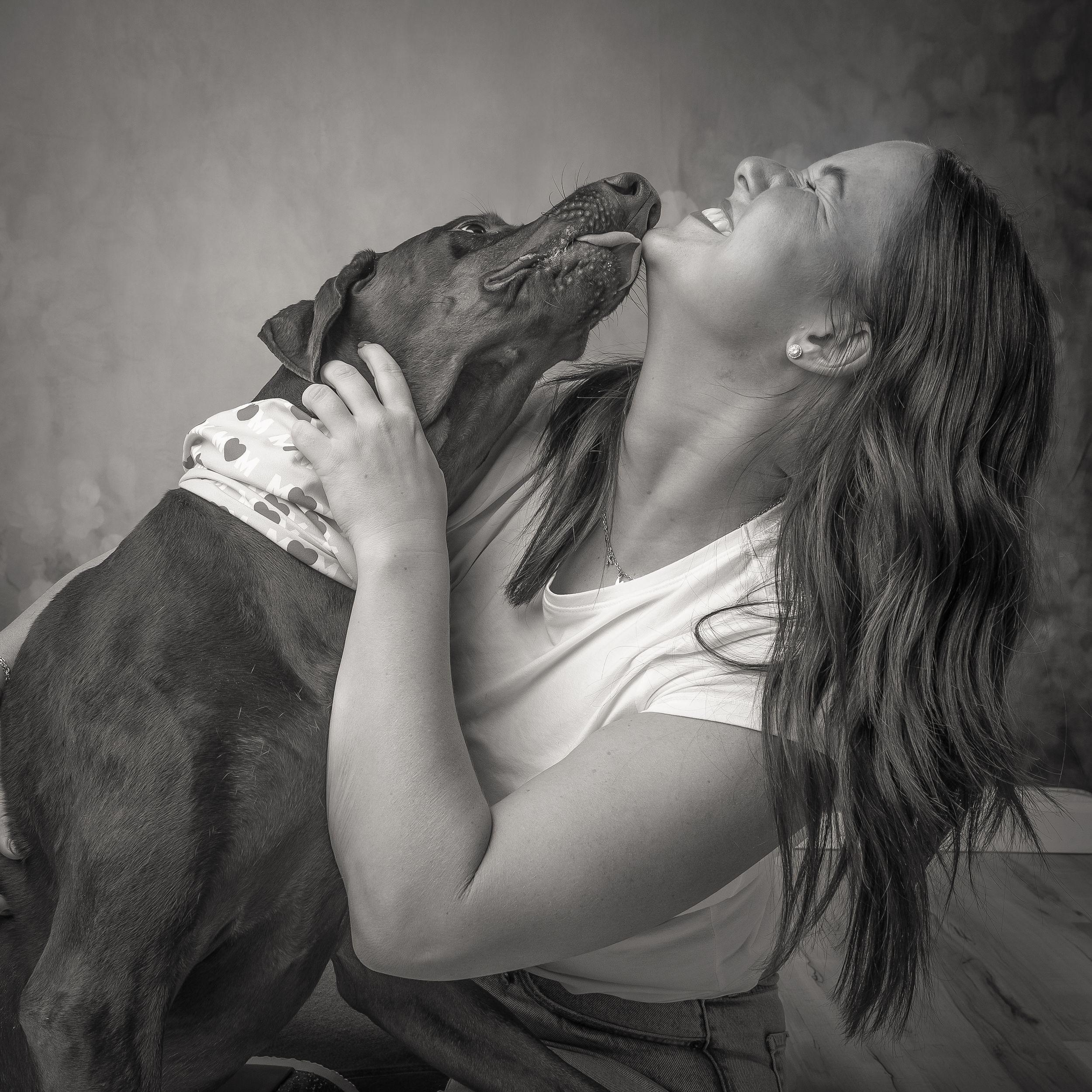 Black and white photo of a joyful woman laughing as her Doberman dog licks her face in a loving moment.