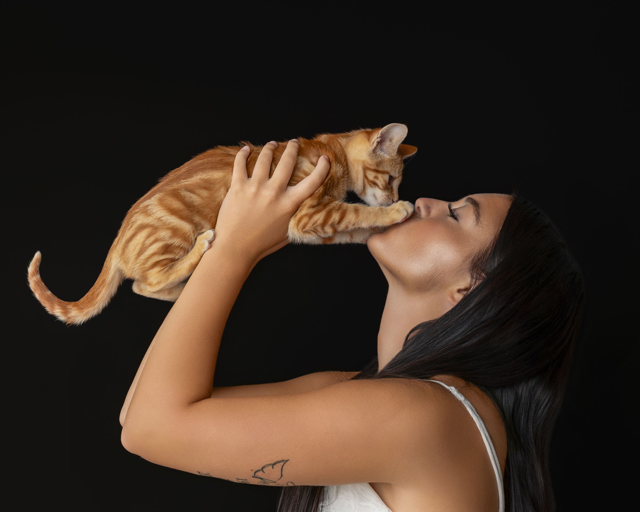 A young woman with dark hair lifts an orange tabby cat to her face for a kiss against a black background.