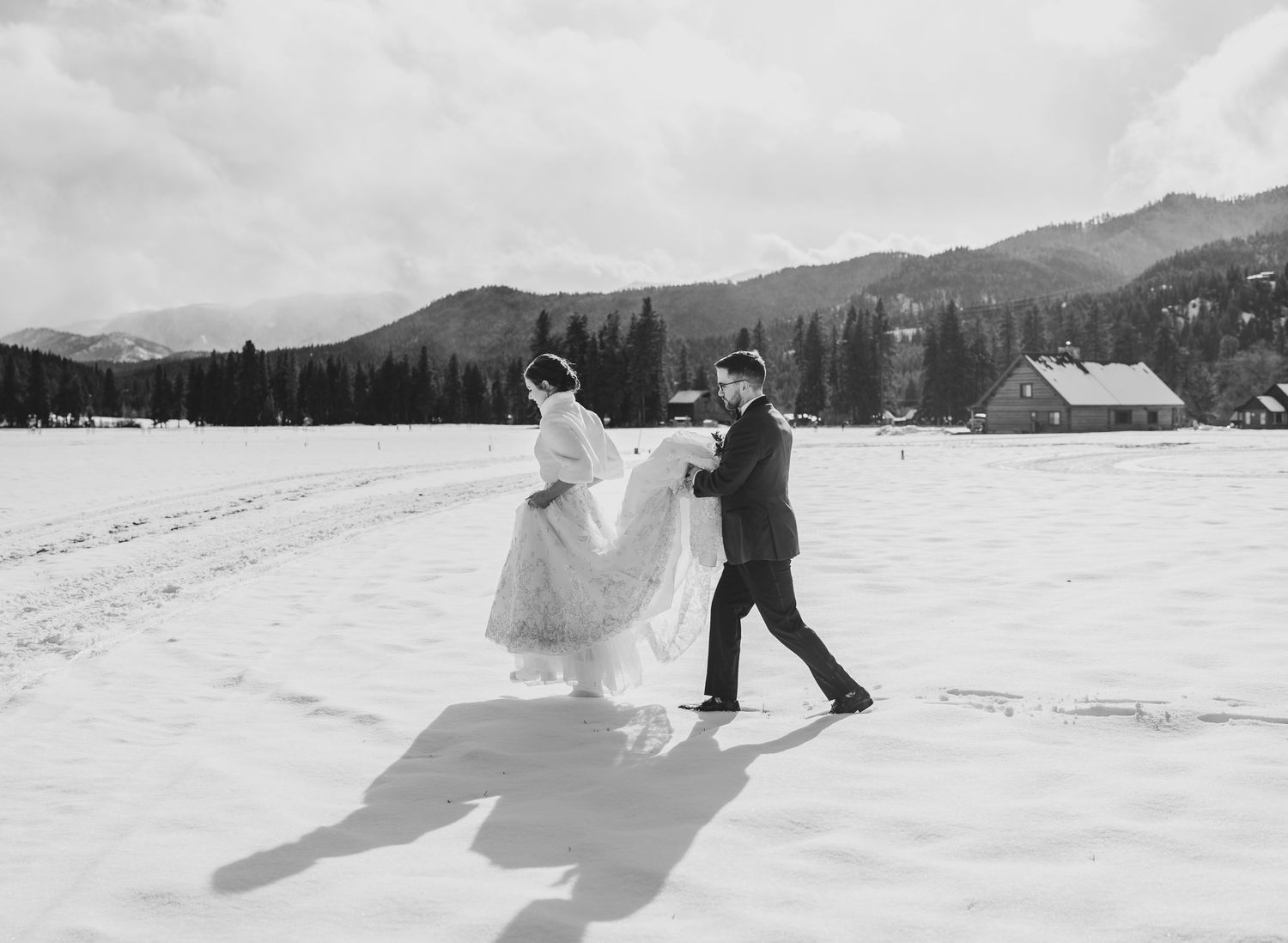 A couple dancing in the snow with mountain scenery and a cabin in the background.