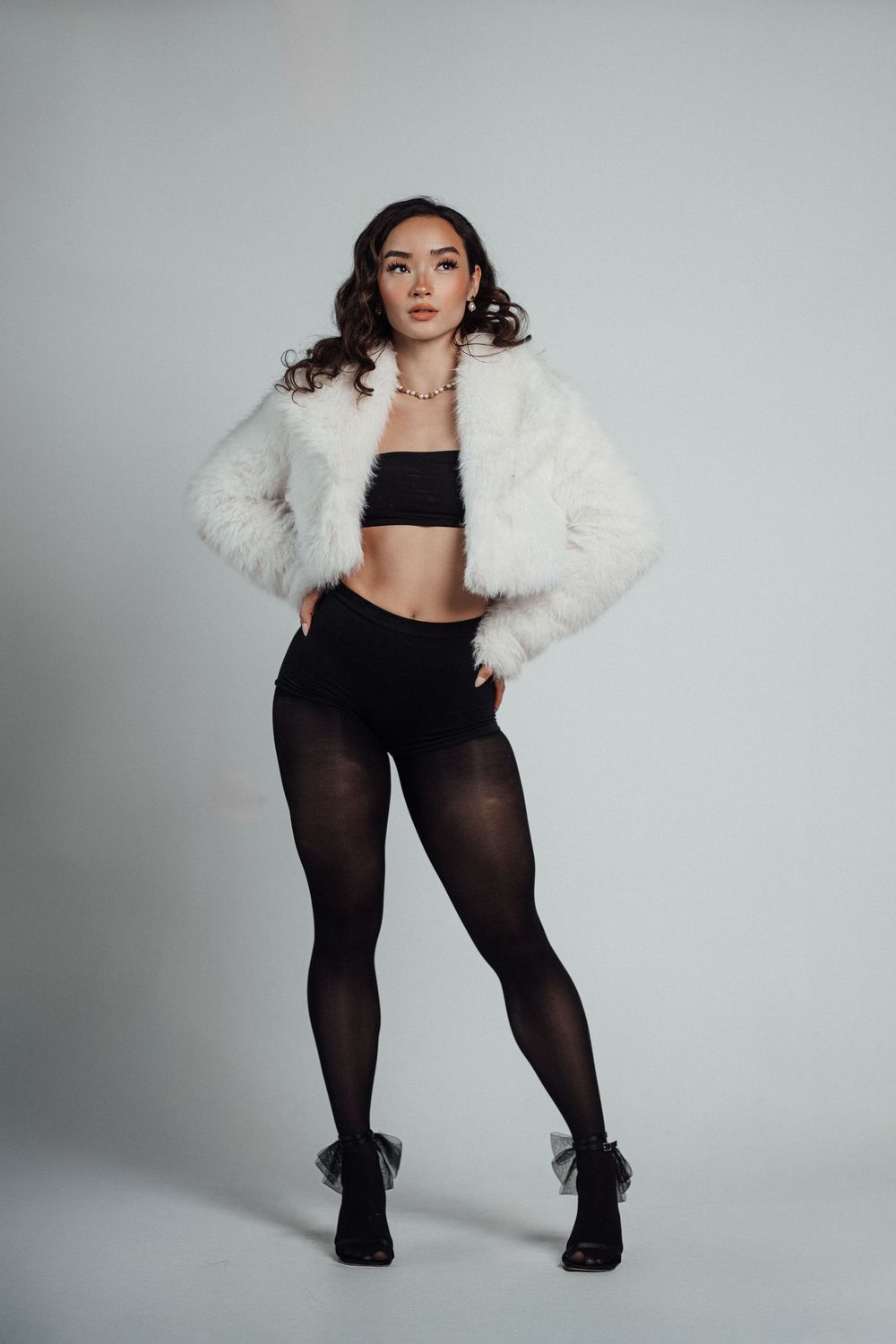 Woman posing in white faux fur jacket, black crop top, sheer tights, and ankle boots on light background.